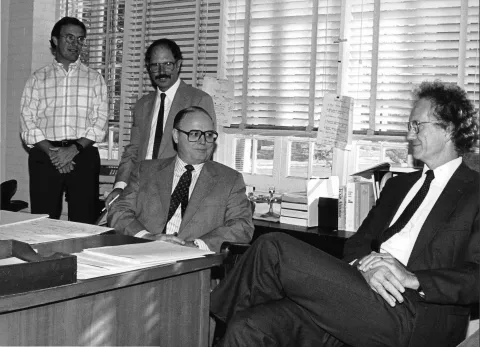 This image shows Chris King, Bob Glover, Ray Marshall, and Bob McPherson during a meeting in the 1970s.  It is a black and white picture and shows two men standing and two men sitting.  All four are in business attire.