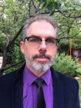 This is a closeup picture of Greg Cumpton outdoors.  He has grey hair with a beard and is wearing black framed glasses.  He is wearing a dark blue suit with a purple shirt and black tie.