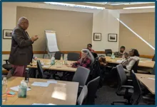 This is a picture of a presentation at the Opportunity Youth Forum. It shows a man standing at the front of the room with people seated at several tablesin a classroom-style setting.