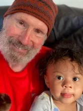 This is an image of Bob Glover and his grandchild. Bob has a gray beard and is wearing a red shirt and brown beanie, and his grandchild has dark curly hair and is wearing a light-colored onesie.  Both are smiling.