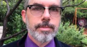 This is a closeup picture of Greg Cumpton outdoors.  He has grey hair with a beard and is wearing black framed glasses.  He is wearing a dark blue suit with a purple shirt and black tie.