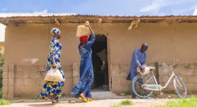 This is the report cover for Nuru Nigeria 2023 Impact Report.  It shows two Nigerian women in local dress carrying sacks of unknown contents walking into a doorway of a building.  There is a Nigerian man, also in local dress, putting a sack on the back rack of his bicycle.
