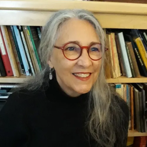 This image is a close-up of Cynthia Juniper, a Research Associate at the Ray Marshall Center at the University of Texas at Austin.  She has a light complexion, gray hair, red glasses, and is smiling.
